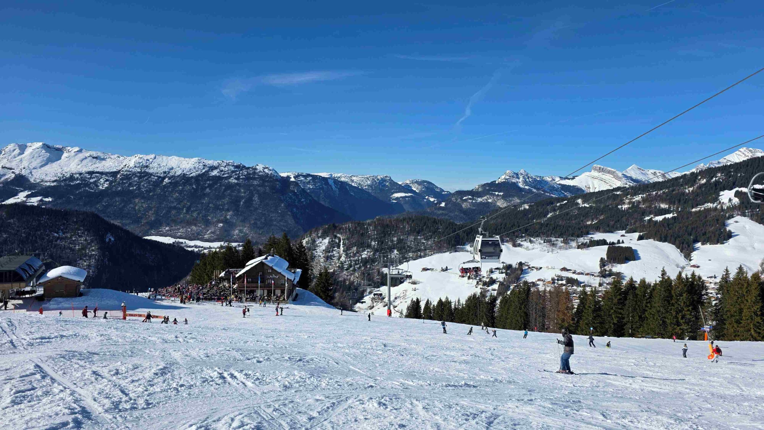 Skiers on a wide blue piste in La Clusaz with the Crêt du Merle gondola and a mountain restaurant in the background"