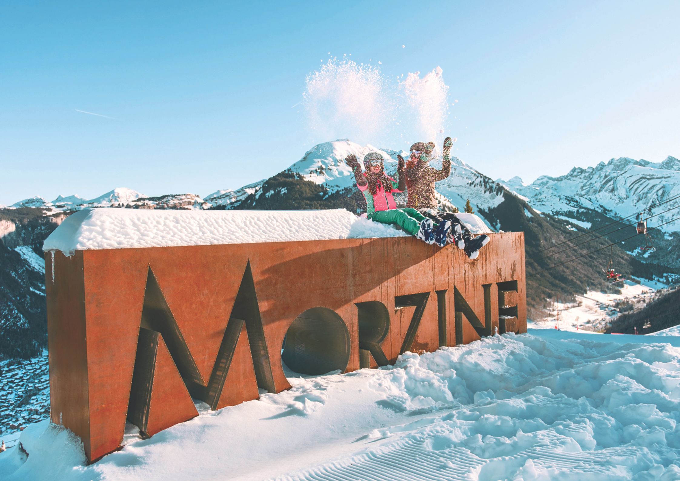 Skiers throwing snow behind the iconic large metal Morzine sign with alpine mountains in the background.