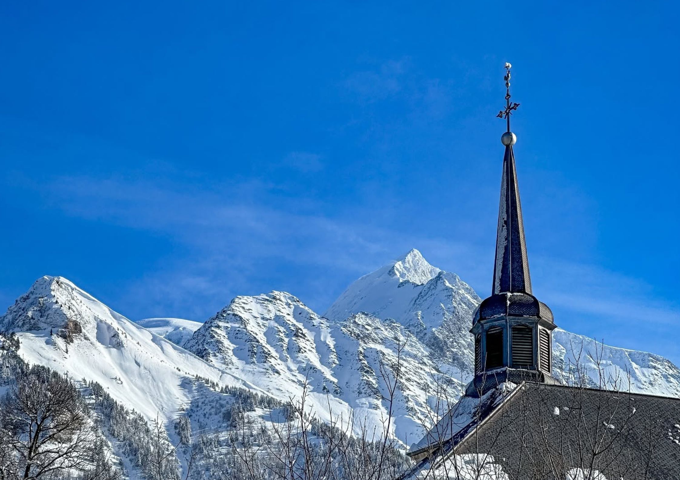 Close-up of a traditional historic church steeple in Saint-Gervais-les-Bains with snow-covered Mont Blanc peaks under a clear blue sky.