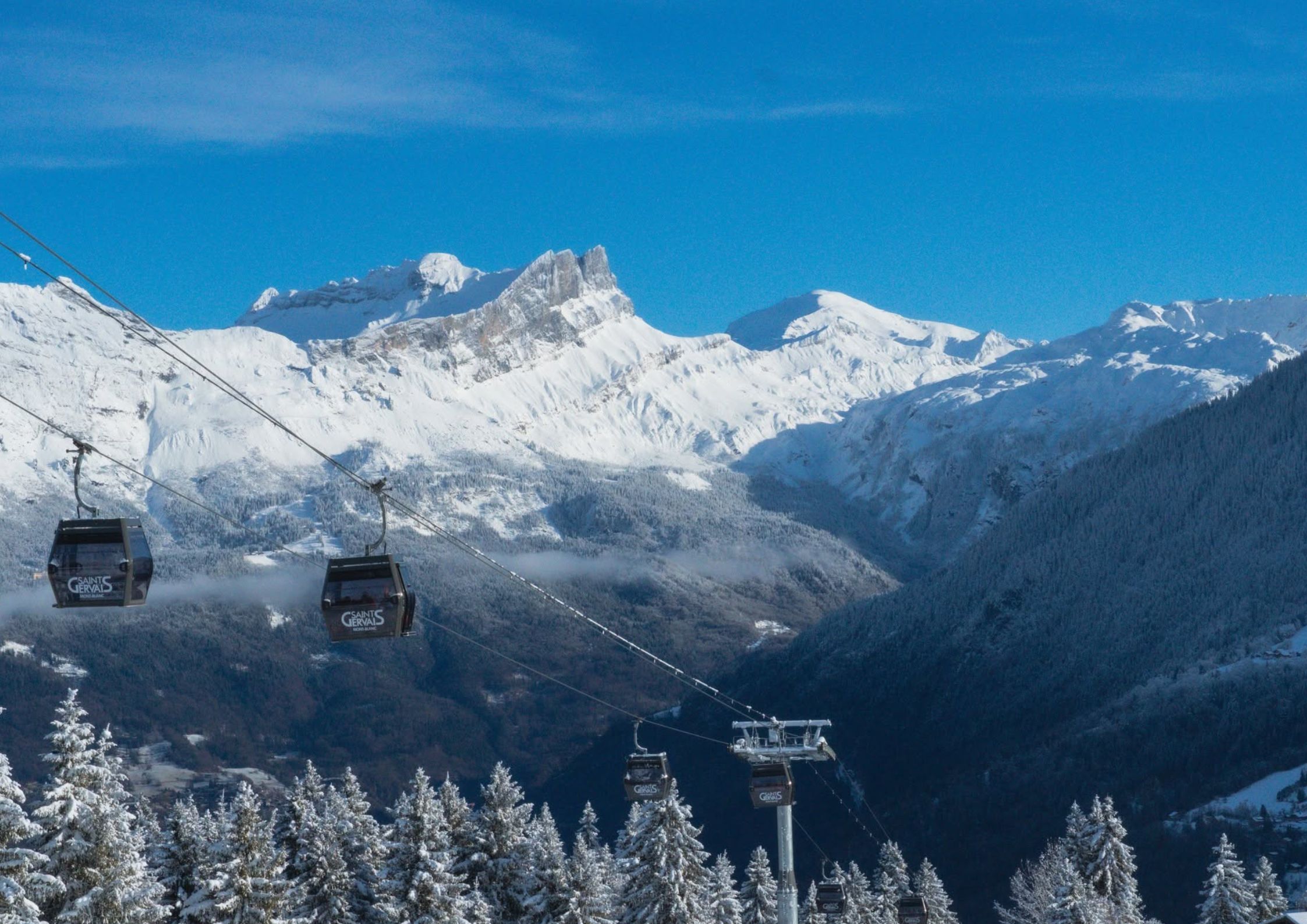 Saint Gervais Mont-Blanc gondola cabins passing over snow-laden pine trees with a dramatic alpine mountain range in the background.
